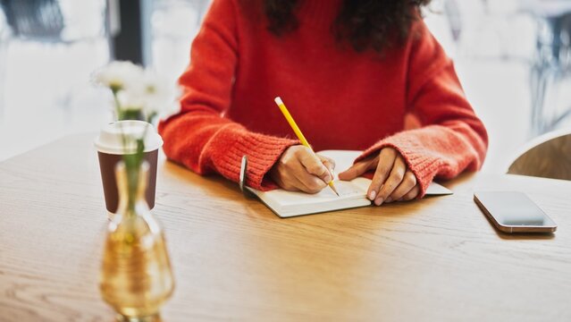 Close up of woman in red sweater writing notes with pencil in notebook at cafe table, takeaway coffee cup nearby. Female with curly hair studying or journaling indoor, smartphone placed on wooden desk