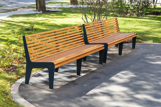 Two modern wooden benches with black metal frames in a park with green grass and trees, placed on a paved surface, surrounded by nature