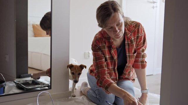 Woman paints door frame with small roller beside paint tray on protective sheet, with dog nearby. Concept of renovation, DIY and wall painting