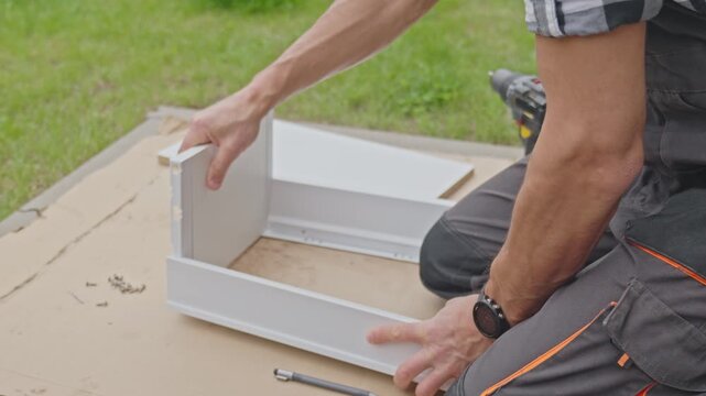 Man fits drawer panels together on cardboard near grass. Concept of DIY furniture assembly, cabinet installation and repair