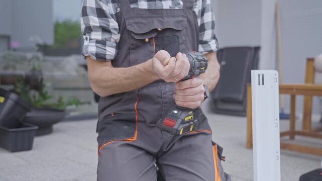 Man changes drill bit on cordless drill beside drawer panel at furniture assembly workspace. Concept of DIY repair and tool preparation