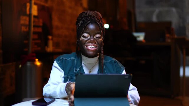 Overjoyed young woman vitiligo skin waving hand digital tablet online video chat outdoor caf&eacute; terrace. Happy female user laughing genuinely live streaming social media app casual content
