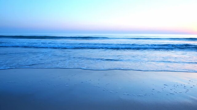 Gentle ocean waves lap onto a serene beach at dawn. The soft pastel sky reflects beautifully on the wet sand.