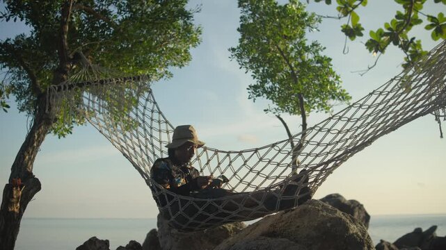 A man wearing sunglasses and a bucket hat reading a book in a hammock on the beach, enjoying a quiet tropical getaway beside the ocean.