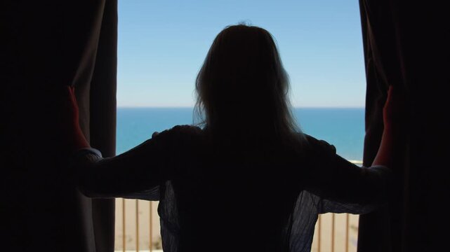 Woman opening curtains in dark room and revealing bright sea view through hotel window during vacation morning by the coast