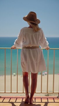 Woman in white dress and straw hat standing on balcony and looking at turquoise sea during relaxing summer beach vacation