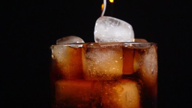 Pouring Dark Soda Drink into Glass Full of Ice Cubes on Black Background. Cola Beverage Over Ice Cubes in Studio. Clear Ice Cubes for Cold Summer Drink.
