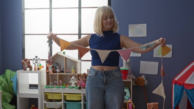 Woman raises arms holding colorful bunting with exposed midriff while smiling in a preschool classroom with toys and storage bins; playful joy.