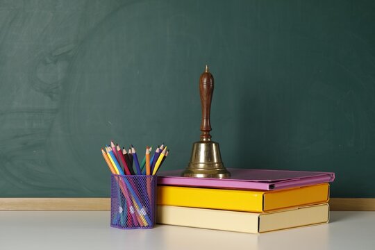 School golden bell, folders and colorful pencils in holder on white table near green chalkboard