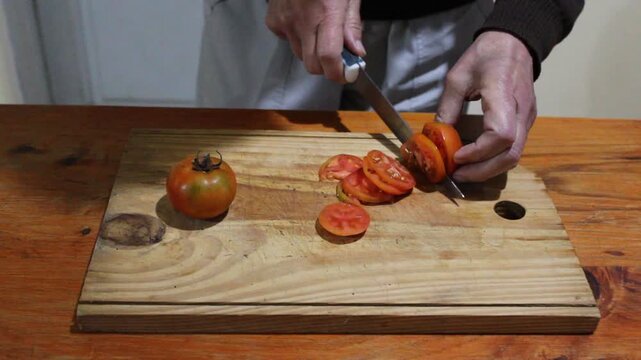 Persona cortando tomates org&aacute;nicos en rodajas con una hoja afilada sobre una tabla de madera r&uacute;stica en la cocina. La escena transmite la cocina casera, la alimentaci&oacute;n saludable y preparaci&oacute;n.