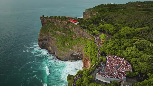 Uluwatu Temple perched on a dramatic Bukit Peninsula sea cliff, crashing waves and a packed outdoor amphitheater where spectators await the traditional Kecak fire dance performance. Aerial view shot
