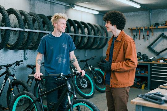 Young adult men discussing bicycle repair in workshop, examining mountain bike and service options. Useful for cycling retail, maintenance service, small business marketing