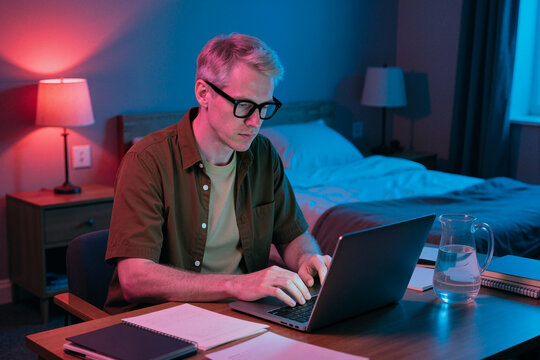 Young man working on laptop at home desk, typing and focusing on remote tasks in bedroom. Useful for remote work, freelance business, productivity marketing