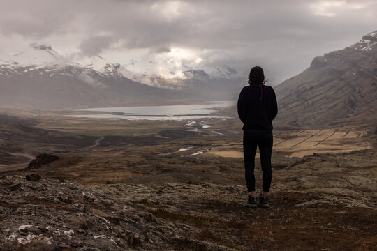 Voyageuse contemplant un paysage spectaculaire d&rsquo;Islande compos&eacute; d&rsquo;une vall&eacute;e, d&rsquo;un lac et de montagnes partiellement enneig&eacute;es sous une lumi&egrave;re douce et un ciel nuageux.
