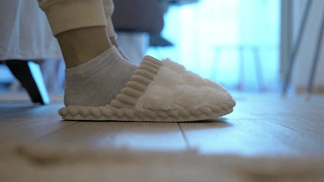 Woman putting on slippers after waking up. Close-up of a woman's feet getting out of bed in the morning, putting on cozy white slippers, and walking away on the wooden floor, starting a new day