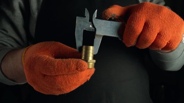 Worker in orange gloves measures brass fittings with caliper tool, showcasing precision in a workshop setting with dark background elements