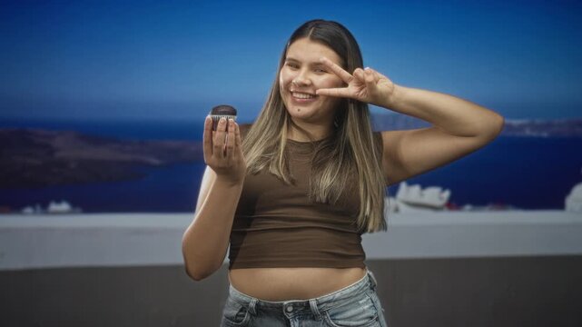 Young woman holds chocolate muffin in one hand, flashes peace sign with other, showing bare midriff on a building terrace overlooking a blue sea at dusk; playful indulgence.