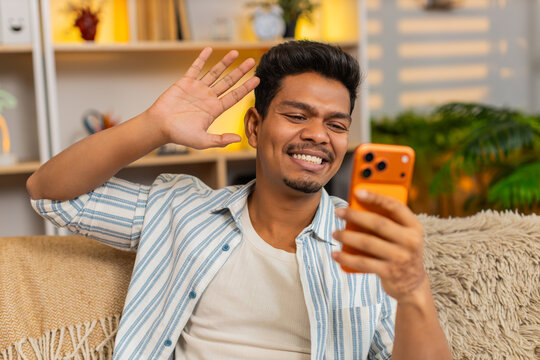 Indian man on home sofa waves at smartphone camera during video call greeting with friendly smile and chat. Arabian guy on home sofa speaks on screen then waves goodbye with warm casual mood indoors.