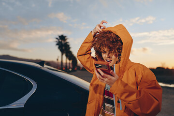 Obraz premium Woman lifestyle portrait, curly hair under hooded orange jacket, smiling, holding phone near car at sunset outdoors, palm trees, warm evening light, casual travel, communication, city road