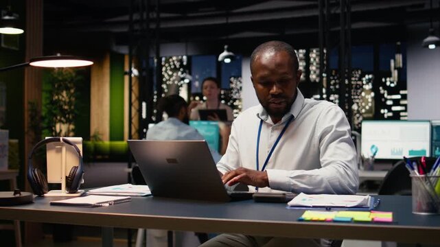 African american worker typing on laptop keyboard at office desk, doing computer tasks and working overnight. Adult man using notebook at work, finishing accounting assignment.