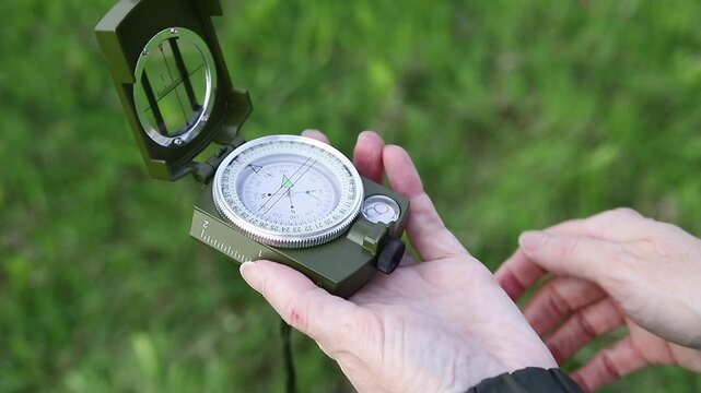 Hand opens the lid of an army compass against a background of green grass. Orienteering with a compass. Concept of travel, adventure, and navigation.