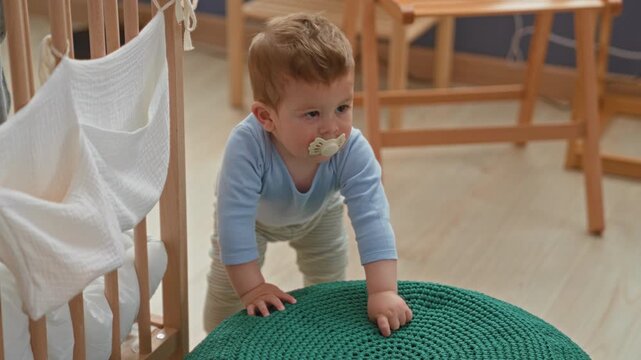 Toddler boy with pacifier standing and leaning on green knitted pouf in building; curiosity and discovery.