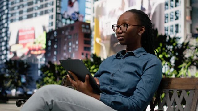 Black digital nomad with tablet, bench in the park downtown, city life relax and productivity with wireless device. Woman ensures task management within urban landscape outdoors, metropolis.