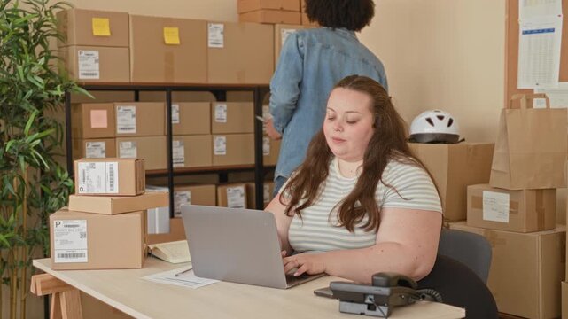 Woman typing on laptop and sorting shipping parcels at a packing desk with stacked boxes and labels in building; entrepreneurship confidence.
