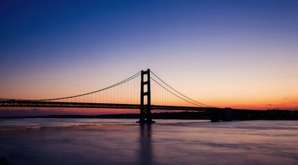 Obraz premium Suspension bridge silhouette at dusk over calm water. Urban skyline fades into twilight sky with orange and blue hues. Majestic engineering marvel over peaceful river.