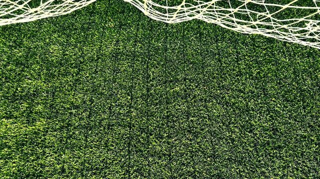High-angle, top-down view of a white soccer goal net blowing gently in the wind, casting a moving grid-pattern shadow onto the vibrant green artificial turf field below.