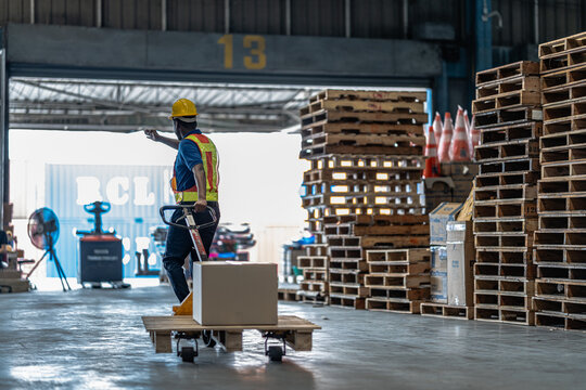A warehouse operator utilizing a manual hydraulic pallet jack to maneuver heavy cargo boxes past stacked wooden pallets inside a commercial logistics distribution terminal.