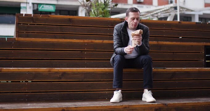 A man sits alone on wooden benches in a bustling urban park. He relaxes while eating a snack under the warm sunlight, surrounded by lively city activity
