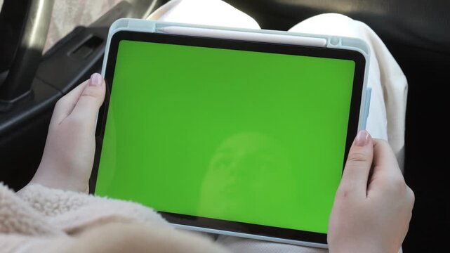 Hands hold tablet with green screen. Person sits comfortably in car. Feet rest on seat beside device. Ideal for digital marketing or travel ads. Green screen allows custom content placement