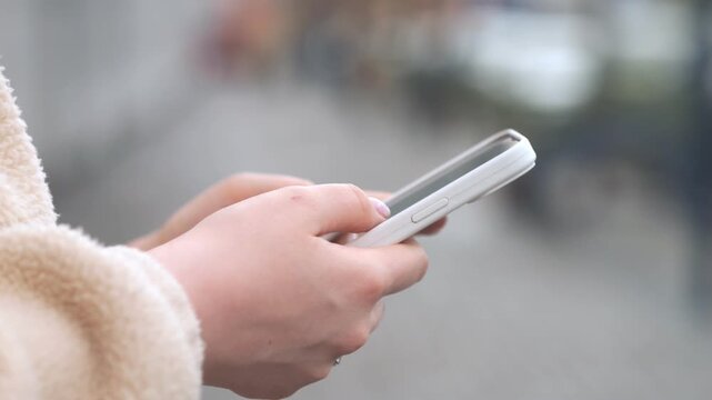 Close-up of hands holding phone outdoors. Fingers scroll on screen. Person wears cozy fleece jacket. Background blurred, suggesting urban street. Ideal for tech, communication, or daily life themes