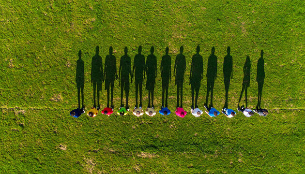 Team formation from above: A group of people stand in a line on a grass field, their shadows stretching long in the sun.