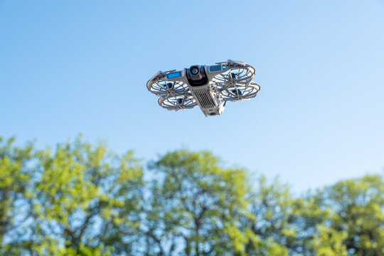 A drone hovering mid-air against a clear blue sky, equipped with a forward-facing camera and propeller guards, with green treetops in the background