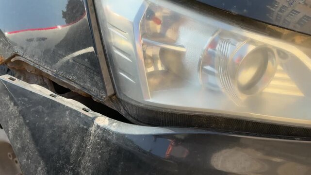 Close-up damaged black car front shows torn bumper and rusted fender, headlight illuminated, steel wheel with scuff marks, gravel