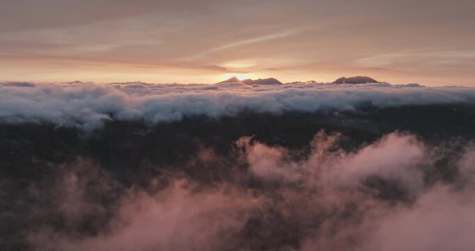 Dramatic aerial view of morning Mount Batur volcano, sunlight breaking through soft pink and orange clouds, revealing mountain peak emerging from atmospheric inversion in Bali. Drone flight footage