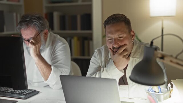 Two men working late at desks in a building, one rubbing his temples and the other with hands clasped over a laptop; overnight fatigue.