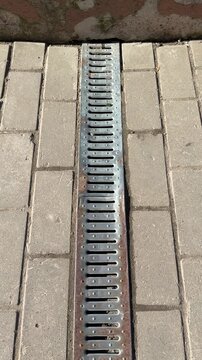 Metal grate spillway on paving stones white downpipe above, patterned metal cover set into paver joints, thin rust streaks