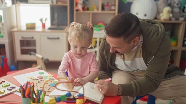 Man leaning over little girl drawing with colored markers and notebook at a small table in building playroom; learning tender bonding.