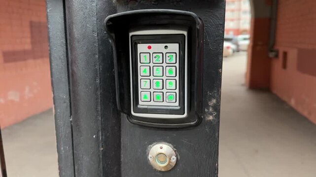 Analog intercom keypad on iron gate, weathered black frame, green backlit numeric buttons, round scanning key below keypad, brick