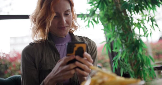 Smiling woman using a mobile phone in a bright Cafe during daylight hours next to plant decor, possibly sharing content or connecting with friends