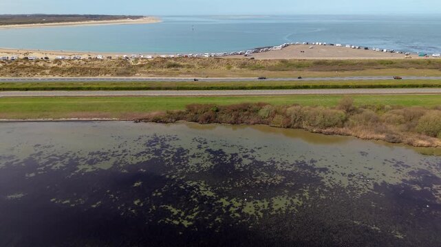 Aerial shot of a coastal landscape featuring a shallow lagoon with dark algae, a parallel highway with moving cars, and a sandy beach parking area filled with numerous campers and RVs by the sea.