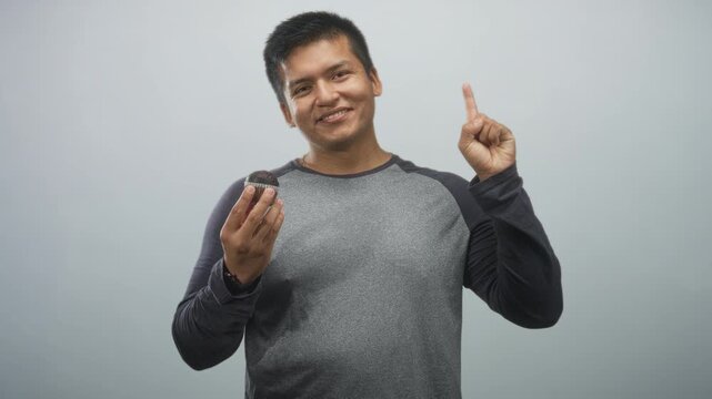 Man holding a chocolate muffin while pointing index finger in studio with plain grey backdrop; cheerful suggestion.