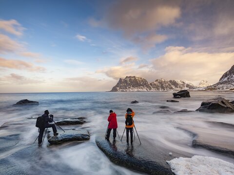 Photographers with tripod on smooth rocks on Utakleiv beach in a dramatic cloudy atmosphere, snow-covered mountains in the background, Vestvagoy Island, Lofoten, Norway