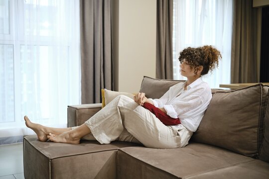 Dreamy woman with curly hair sits comfortably on a beige sofa in a bright living room. She wears a light shirt and relaxed pants while gazing thoughtfully out the window during the day