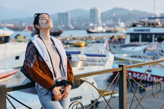 Woman leaning on a railing at a sunny harbor, outdoor fashion in a warm coat, relaxed mood with boats and city skyline in the background, waterfront leisure scene