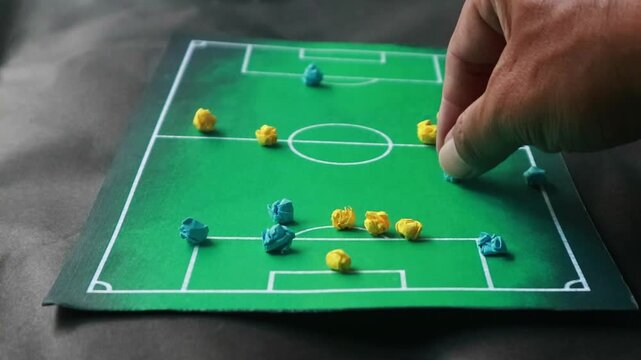 Close-up of a hand moving a mini soccer player on a tactical board with a miniaturized soccer field and sport fans cheering in the background