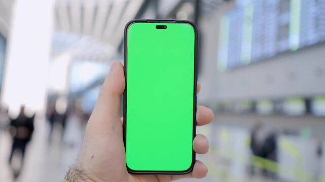 Hand holds phone with green screen. Blurred airport background visible. Phone shows empty green display. Ideal for travel app or digital content mockups. Natural lighting and focused hand detail